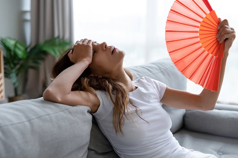 woman sitting on her bed fanning herself with her hand over her forehead.