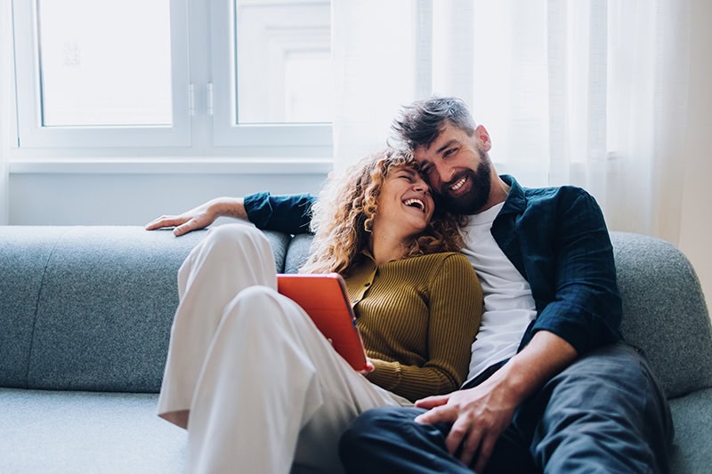 A young couple sitting closely together and laughing on a grey sofa in a brightly lit room.