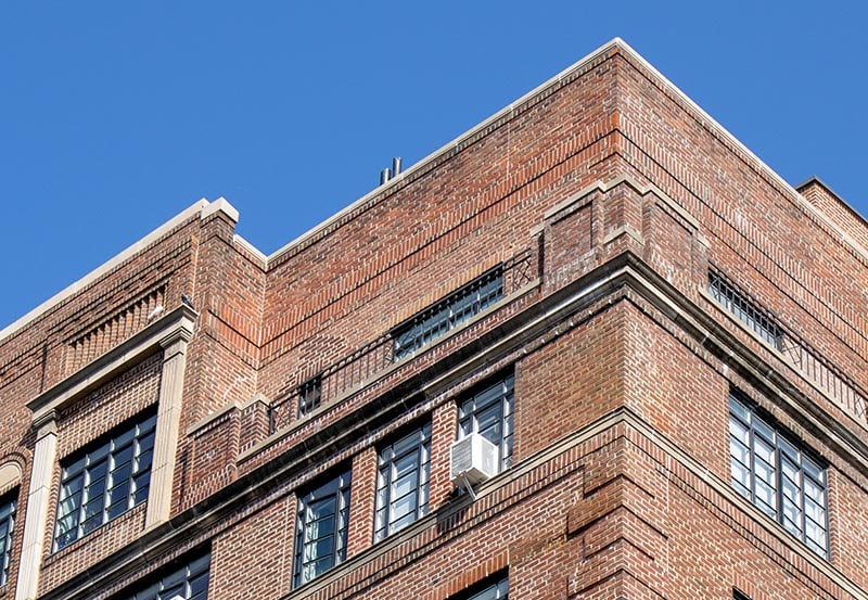 A low-angle view of the upper floors and corner of a classic multi-story brick building against a clear blue sky.