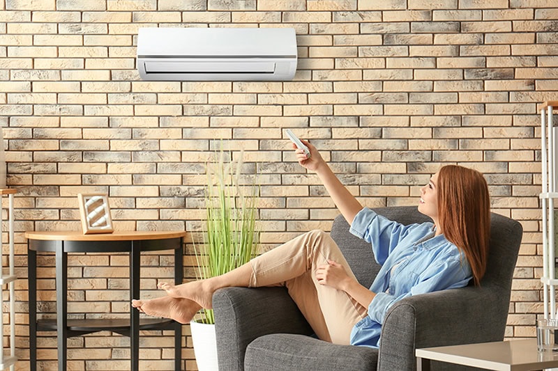 Why Are They Called Ductless Mini Splits? Young woman switching on air conditioner while sitting in armchair at home.